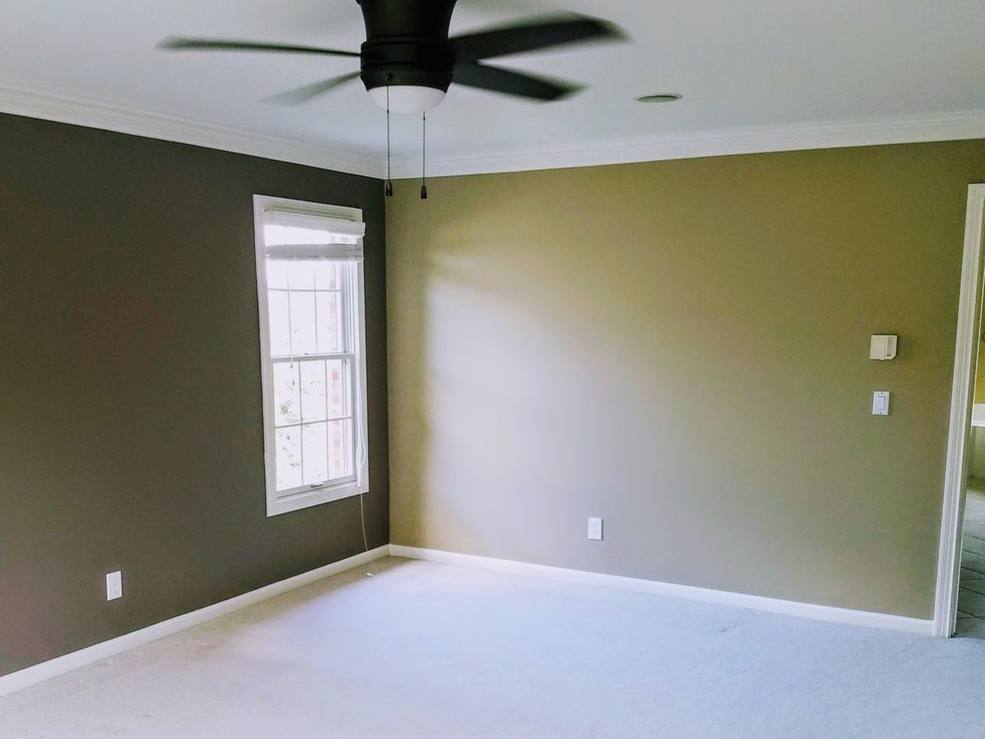 An empty bedroom painted in a deep olive green, with white trim, a ceiling fan and a bright window.